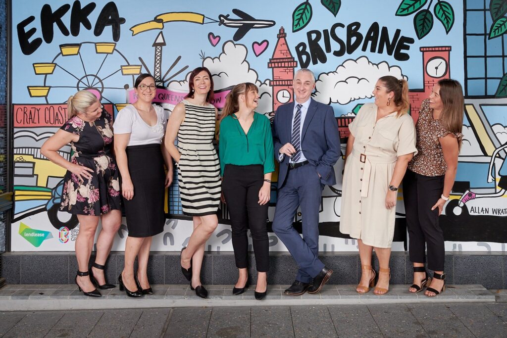A diverse group of smiling professionals posing in front of a colorful Brisbane Ekka-themed mural | MartyK Photography