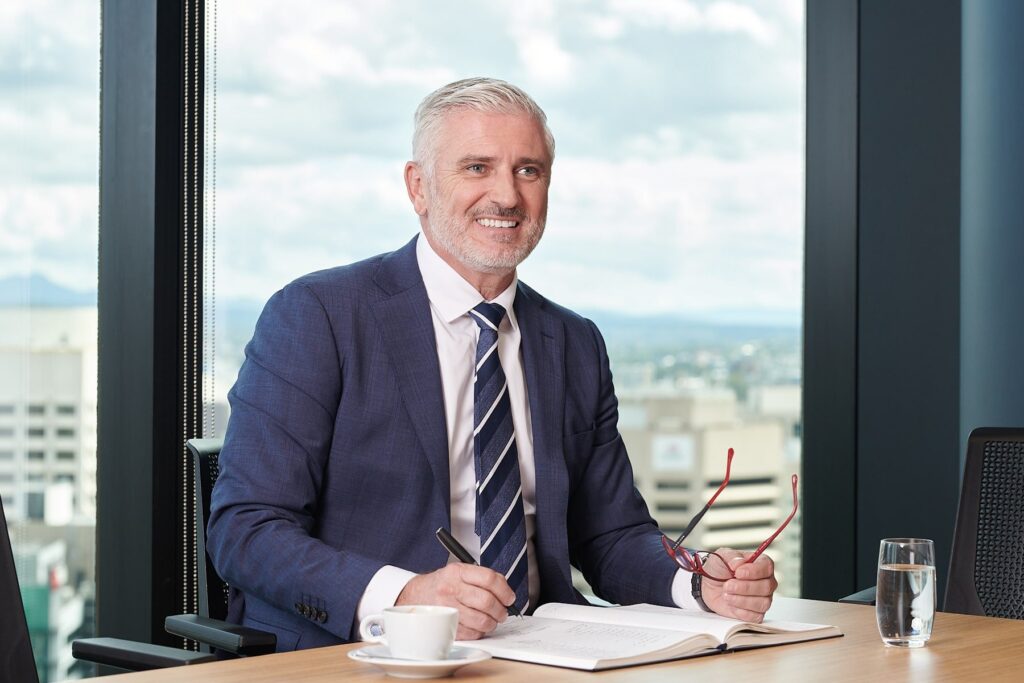 Smiling mature businessman in a blue suit sitting at a desk with a notebook and coffee near a window | MartyK Photography