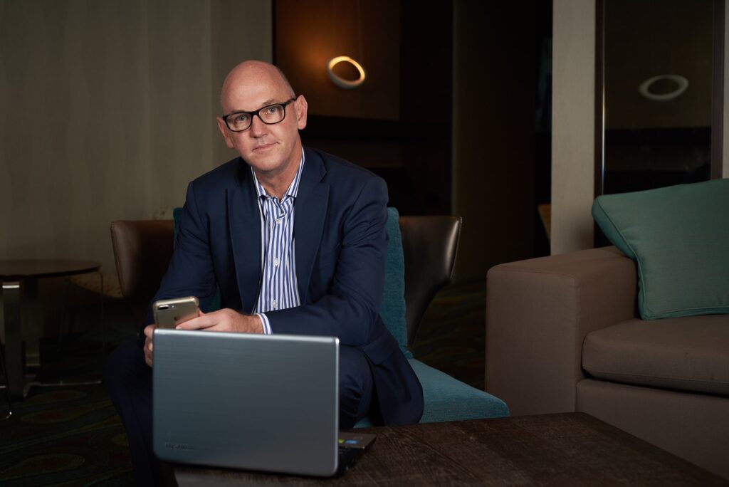 Bald man in a blue suit wearing glasses, using a laptop and smartphone in a modern lounge | MartyK Photography