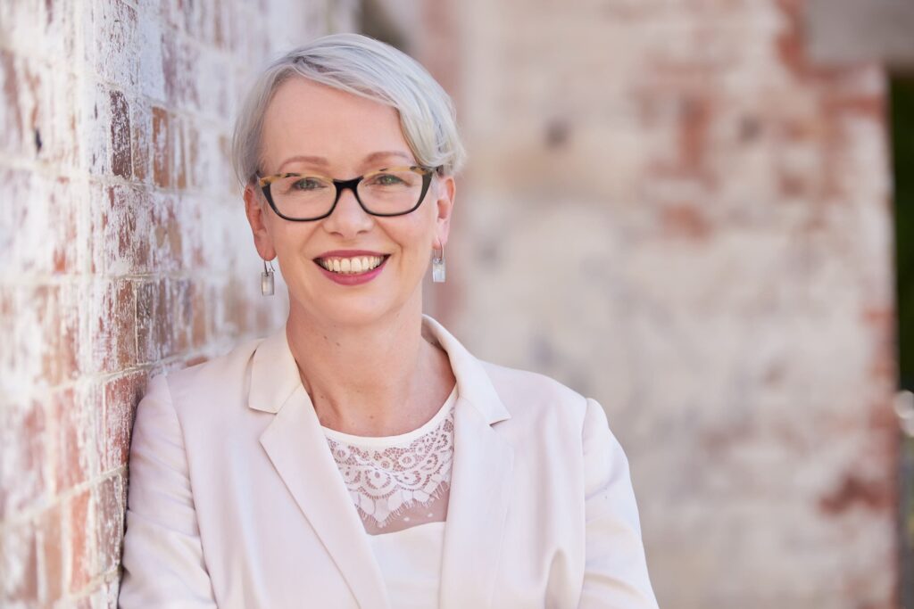 Smiling woman with short grey hair and glasses wearing a white blazer, leaning against a brick wall | MartyK Photography