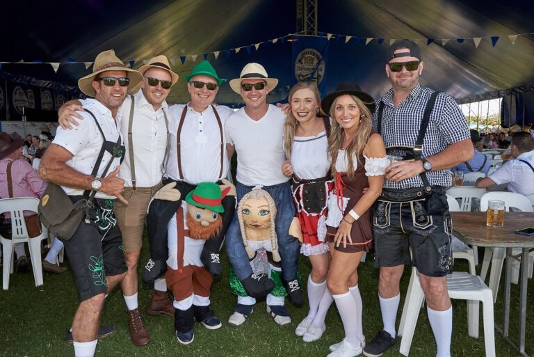 Group of people celebrating Oktoberfest wearing traditional Bavarian lederhosen and dirndls under a blue festival tent | MartyK Photography