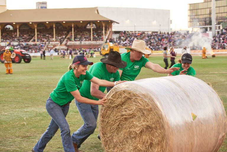 Four people in green shirts pushing a large round hay bale during a fun outdoor community event | MartyK Photography
