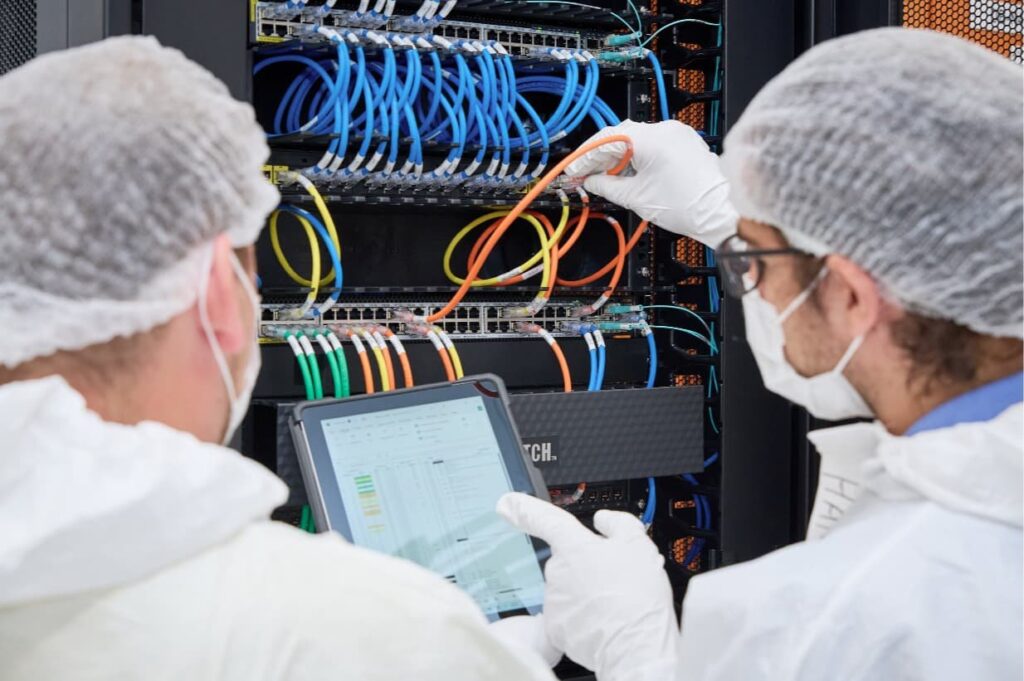 Two technicians in cleanroom suits inspecting server rack network cables and monitoring data on a digital tablet | MartyK Photography